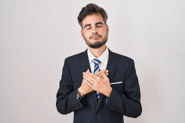 Young hispanic man with tattoos wearing business suit and tie smiling with hands on chest with closed eyes and grateful gesture on face. health concept.