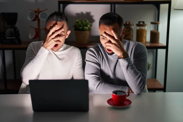Homosexual couple using computer laptop peeking in shock covering face and eyes with hand, looking through fingers with embarrassed expression.