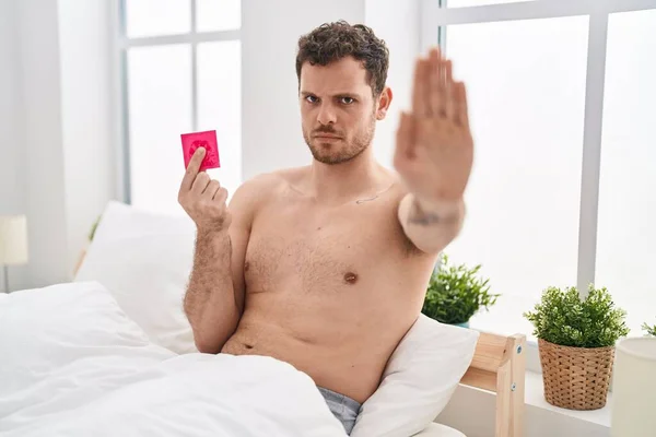 Young hispanic man holding condom in the bed with open hand doing stop sign with serious and confident expression, defense gesture