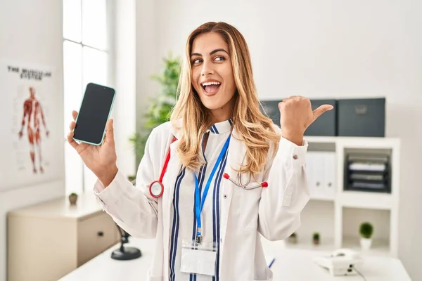 Young blonde doctor woman working at the clinic showing smartphone screen pointing thumb up to the side smiling happy with open mouth