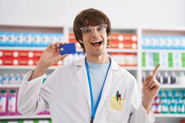 Hispanic young man working at pharmacy drugstore holding credit card smiling happy pointing with hand and finger to the side