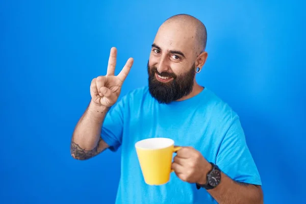 Young hispanic man with beard and tattoos drinking a cup of coffee smiling looking to the camera showing fingers doing victory sign. number two.