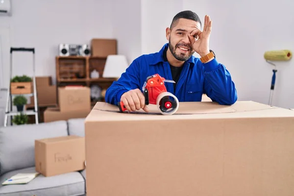 Hispanic man working on moving holding packing tape smiling happy doing ok sign with hand on eye looking through fingers