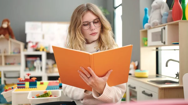 Young Blonde Woman Preschool Teacher Reading Book Relaxed Expression Kindergarten —  Fotos de Stock
