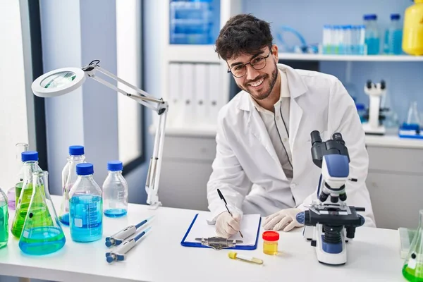 Young hispanic man scientist writing on document holding urine test tube at laboratory