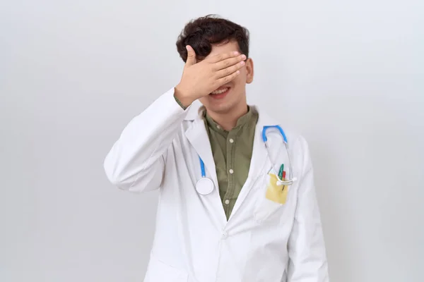 Young non binary man wearing doctor uniform and stethoscope smiling and laughing with hand on face covering eyes for surprise. blind concept.