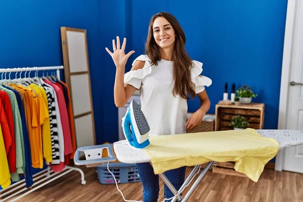 Young hispanic woman ironing clothes at laundry room showing and pointing up with fingers number five while smiling confident and happy. 