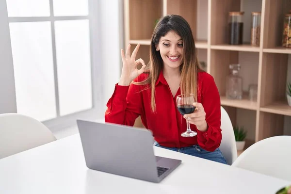 Young brunette woman doing video call drinking red wine doing ok sign with fingers, smiling friendly gesturing excellent symbol