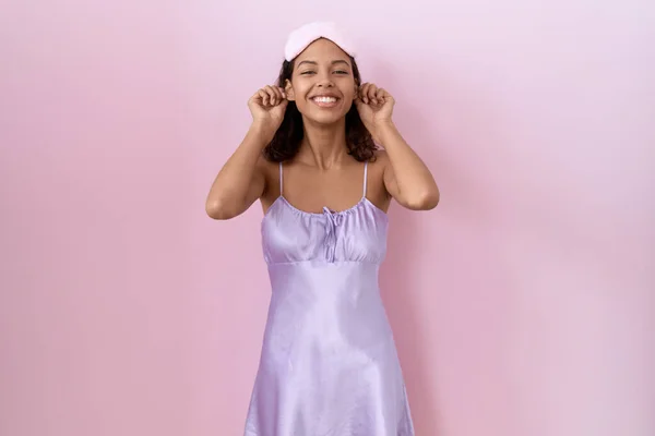 Young hispanic woman wearing sleep mask and nightgown smiling pulling ears with fingers, funny gesture. audition problem