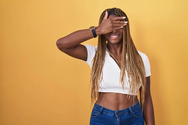African american woman with braided hair standing over yellow background smiling and laughing with hand on face covering eyes for surprise. blind concept.