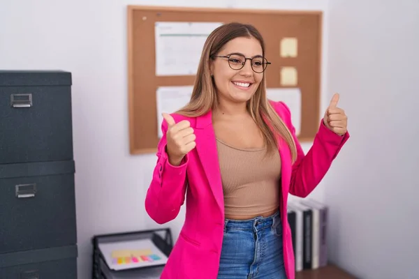 Young blonde woman standing at the office success sign doing positive gesture with hand, thumbs up smiling and happy. cheerful expression and winner gesture.