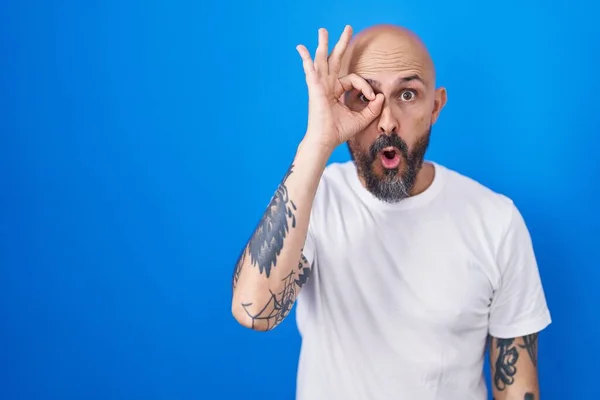 Hispanic man with tattoos standing over blue background doing ok gesture shocked with surprised face, eye looking through fingers. unbelieving expression.