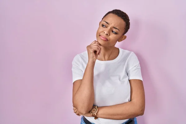 Beautiful african american woman standing over pink background with hand on chin thinking about question, pensive expression. smiling with thoughtful face. doubt concept.
