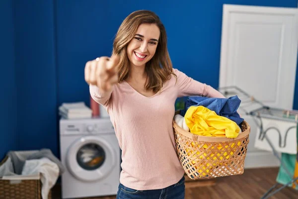 Young woman holding laundry basket pointing to you and the camera with fingers, smiling positive and cheerful