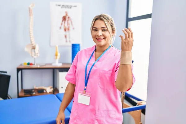 Young hispanic woman working at pain recovery clinic wearing wristband looking positive and happy standing and smiling with a confident smile showing teeth