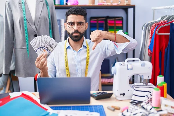 Hispanic man with beard dressmaker designer holding dollars with angry face, negative sign showing dislike with thumbs down, rejection concept