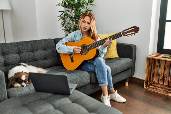 Young caucasian woman having online classical guitar class sitting on sofa with dog at home