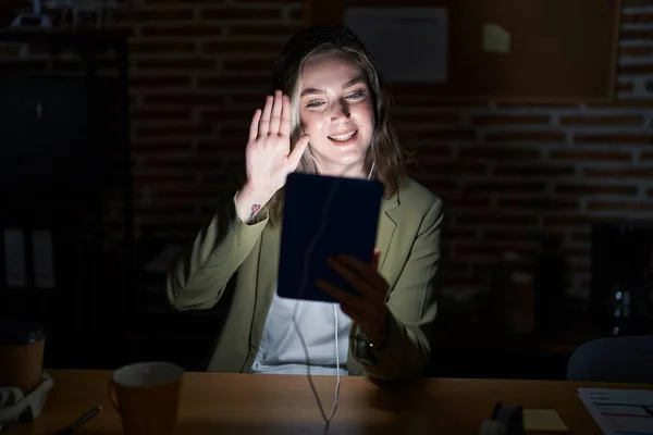 Blonde caucasian woman working at the office at night waiving saying hello happy and smiling, friendly welcome gesture