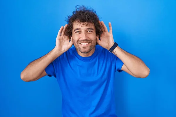 西班牙裔年轻人站在蓝色的背景上 试图听到两只手对著耳朵的手势 好奇地闲聊 听力问题 — 图库照片