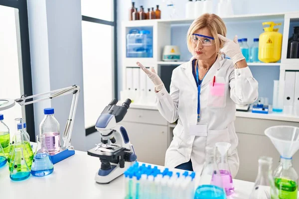 Middle age blonde woman working at scientist laboratory confused and annoyed with open palm showing copy space and pointing finger to forehead. think about it.