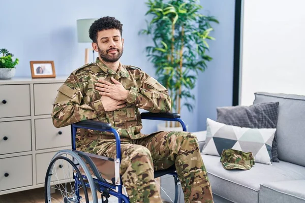 Arab man wearing camouflage army uniform sitting on wheelchair smiling with hands on chest with closed eyes and grateful gesture on face. health concept.
