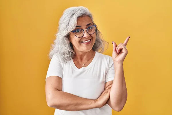Middle age woman with grey hair standing over yellow background with a big smile on face, pointing with hand and finger to the side looking at the camera.