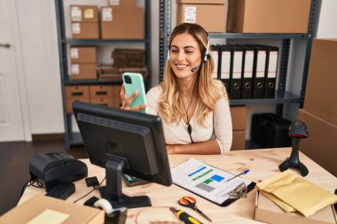 Young blonde woman working at small business ecommerce using headset looking positive and happy standing and smiling with a confident smile showing teeth 