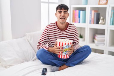 Young hispanic man eating popcorn sitting on the bed watching a movie smiling and laughing hard out loud because funny crazy joke. 