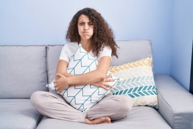Hispanic woman with curly hair sitting on the sofa at home puffing cheeks with funny face. mouth inflated with air, catching air. 