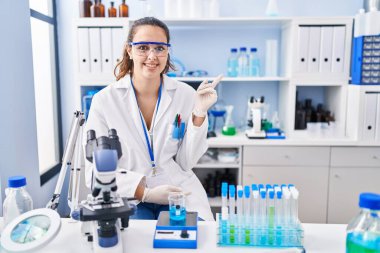Young hispanic woman working at scientist laboratory with a big smile on face, pointing with hand and finger to the side looking at the camera. 