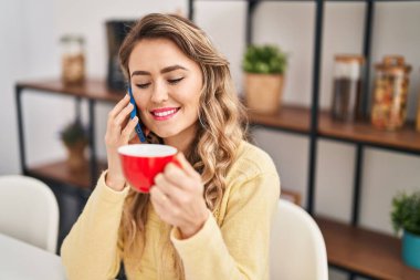 Young woman drinking coffee talking on smartphone at home
