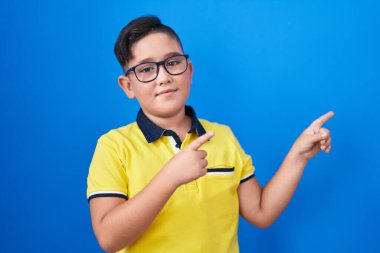 Young hispanic kid standing over blue background smiling and looking at the camera pointing with two hands and fingers to the side. 