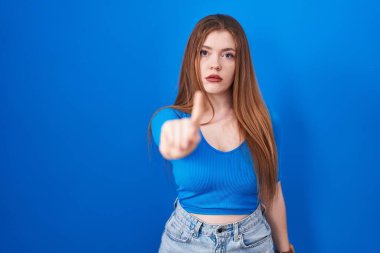 Redhead woman standing over blue background pointing with finger up and angry expression, showing no gesture 
