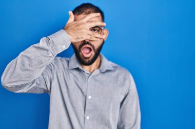 Middle east man with beard standing over blue background peeking in shock covering face and eyes with hand, looking through fingers with embarrassed expression. 