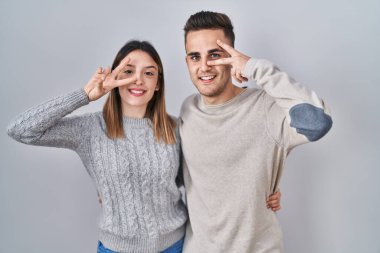 Young hispanic couple standing over white background doing peace symbol with fingers over face, smiling cheerful showing victory 