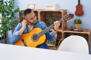 Young hispanic man playing classical guitar sitting on table at home