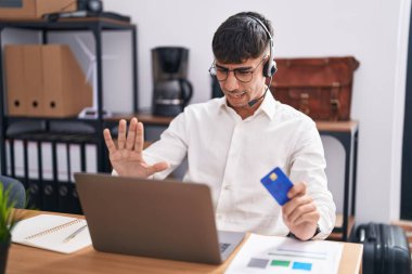 Young hispanic man working using computer laptop holding credit card doing stop gesture with hands palms, angry and frustration expression 