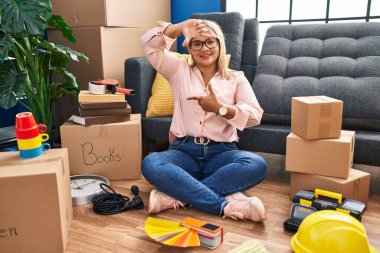 Young hispanic woman moving to a new home sitting on the floor smiling making frame with hands and fingers with happy face. creativity and photography concept. 