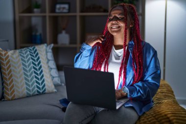 African american woman with braided hair using computer laptop at night smiling confident touching hair with hand up gesture, posing attractive and fashionable 