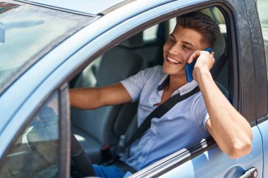 Young hispanic man talking on the smartphone sitting on car at street