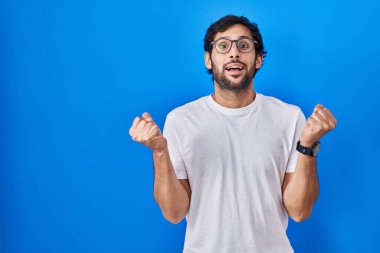 Handsome latin man standing over blue background celebrating surprised and amazed for success with arms raised and open eyes. winner concept. 