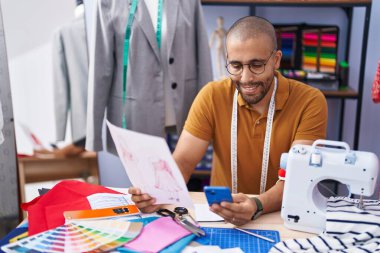 Young latin man tailor using smartphone looking clothing design at atelier