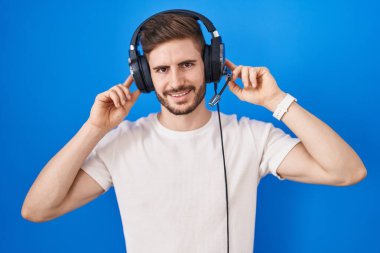 Hispanic man with beard listening to music wearing headphones smiling pulling ears with fingers, funny gesture. audition problem 