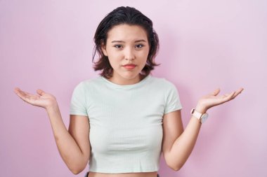 Hispanic young woman standing over pink background clueless and confused expression with arms and hands raised. doubt concept. 