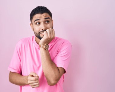 Hispanic young man standing over pink background looking stressed and nervous with hands on mouth biting nails. anxiety problem. 