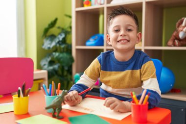 Adorable hispanic boy preschool student sitting on table drawing on paper at kindergarten