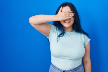 Young modern girl with blue hair standing over blue background smiling and laughing with hand on face covering eyes for surprise. blind concept. 