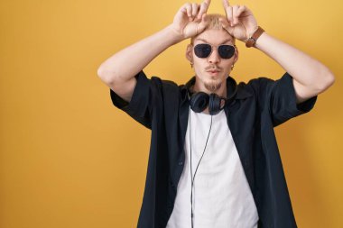 Young caucasian man wearing sunglasses standing over yellow background doing funny gesture with finger over head as bull horns 