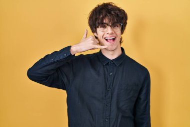 Young man wearing glasses over yellow background smiling doing phone gesture with hand and fingers like talking on the telephone. communicating concepts. 
