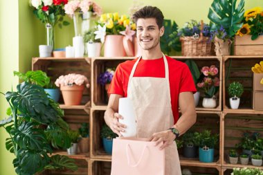 Young hispanic man florist smiling confident putting plant jar on shopping bag at flower shop
